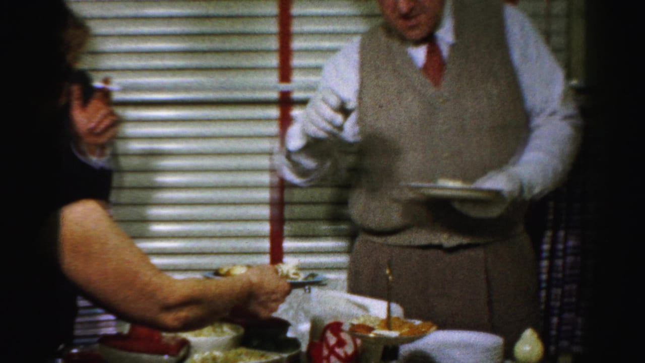 1950s Albany home-movie frame showing adults serving food indoors beside window blinds and a crowded table.