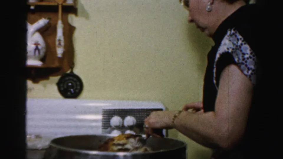 Woman in a patterned-sleeve dress works over a large pot in a mid-century Albany kitchen interior.