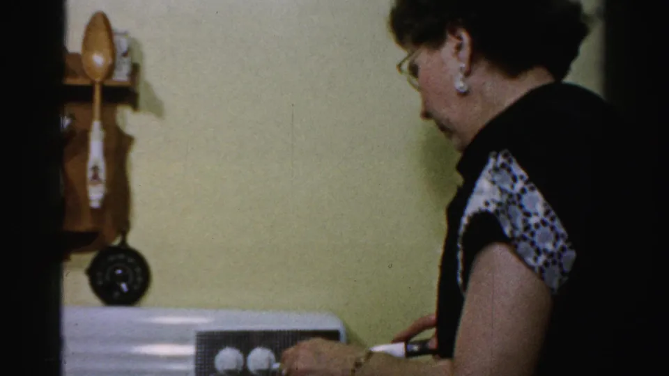Home-movie frame of a woman tending food in a private Albany kitchen, with mid-century fixtures visible behind her.
