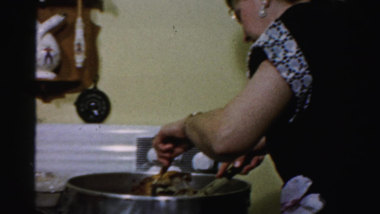 In a 1956 Albany home-movie frame, a woman in glasses leans over a large pan on a white stove while handling food with a fork and knife.