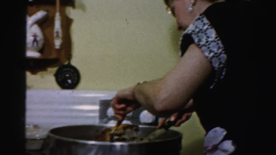 In a 1956 Albany home-movie frame, a woman in glasses leans over a large pan on a white stove while handling food with a fork and knife.