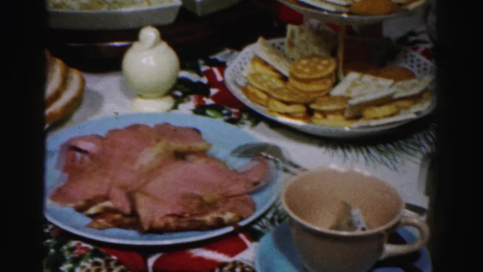 Albany 1956 home-movie frame showing sliced ham on a blue plate, crackers and cheese, a cup and saucer, and festive tablecloth.