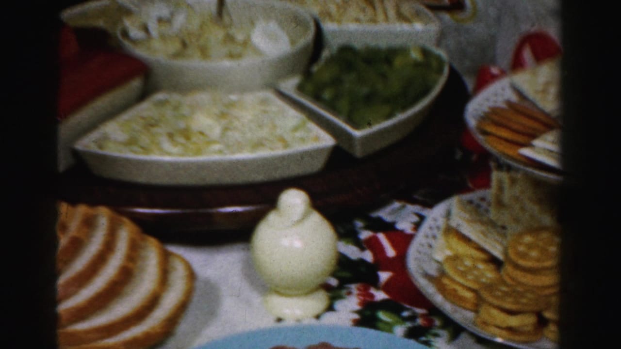 1950s Albany home-movie frame showing a Thanksgiving-style food table with bread, crackers, white serving bowls, and green vegetables.