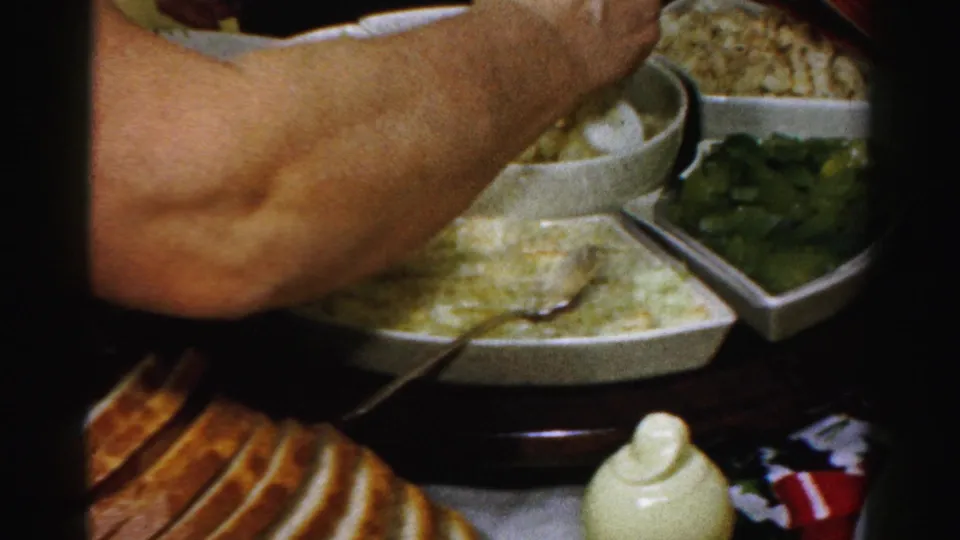 A person's arm reaches across a Thanksgiving food table in Albany, with bread, white bowls, green vegetables, and a serving spoon visible.