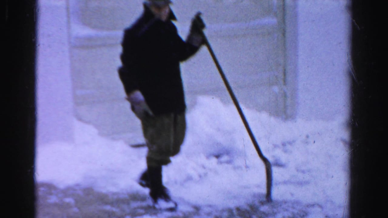 In Albany, a bundled adult man stands in deep snow with a long-handled shovel near a paneled exterior door.
