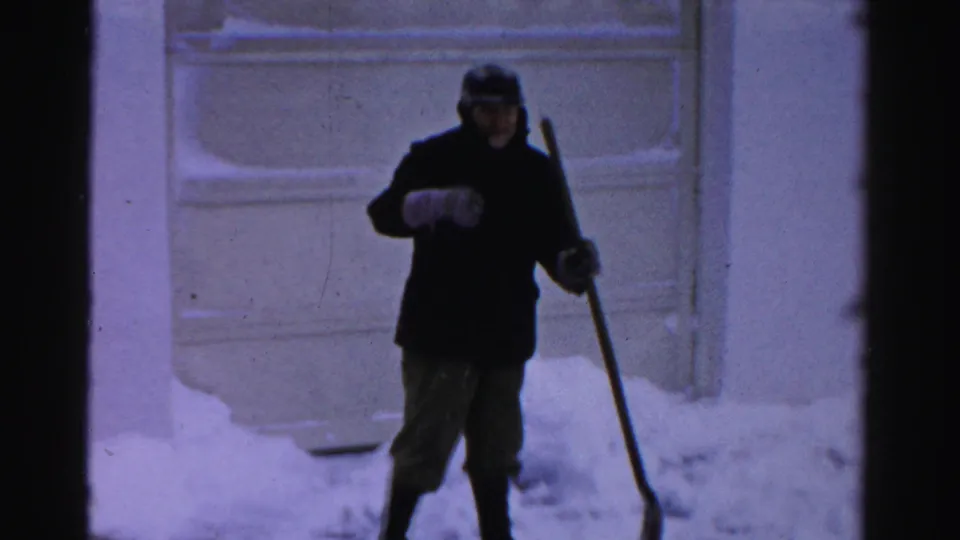 Albany winter home-movie frame showing a bundled man facing the camera while holding a snow shovel near a snow-covered exterior door.