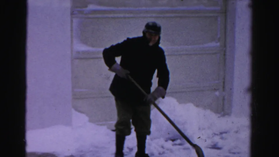 A bundled adult man shovels snow outside a building in Albany, with snow piled around a paneled exterior door.