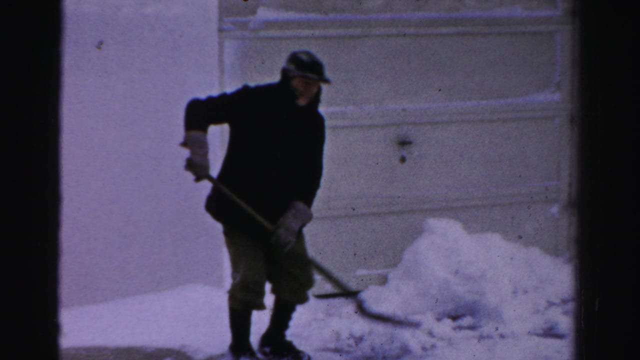In 1956 Albany home-movie footage, a man in a dark coat and cap shovels snow near a residential garage door.
