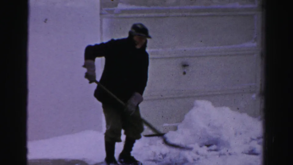 In 1956 Albany home-movie footage, a man in a dark coat and cap shovels snow near a residential garage door.