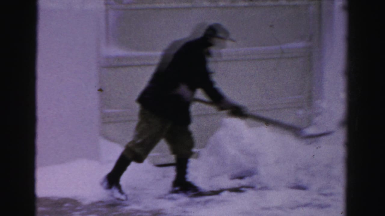 Man bends forward while moving snow with a shovel near a garage.