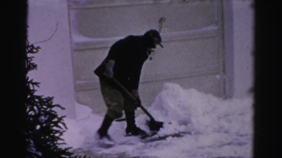 A man in winter clothing clears packed snow from the driveway edge, with the garage door filling the background.