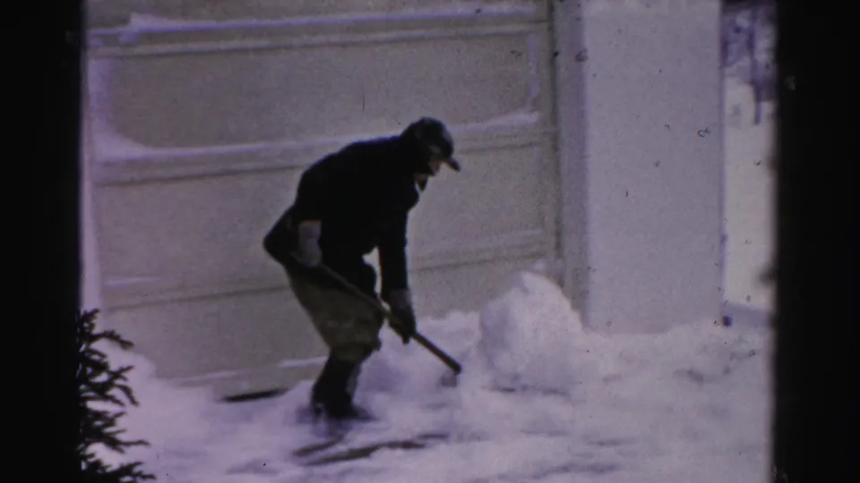 The man pauses or resets his stance with a shovel beside a snow pile in the Albany residential winter scene.