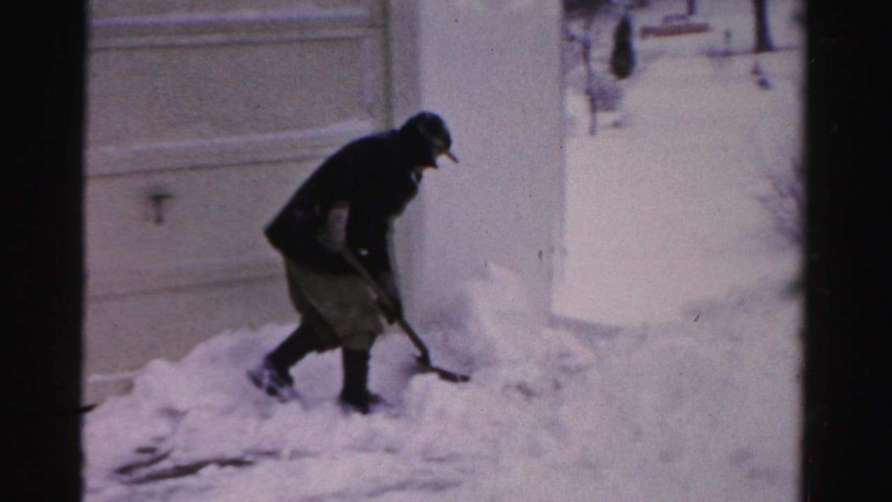 Man in dark winter clothing shovels deep snow beside a garage or house wall.