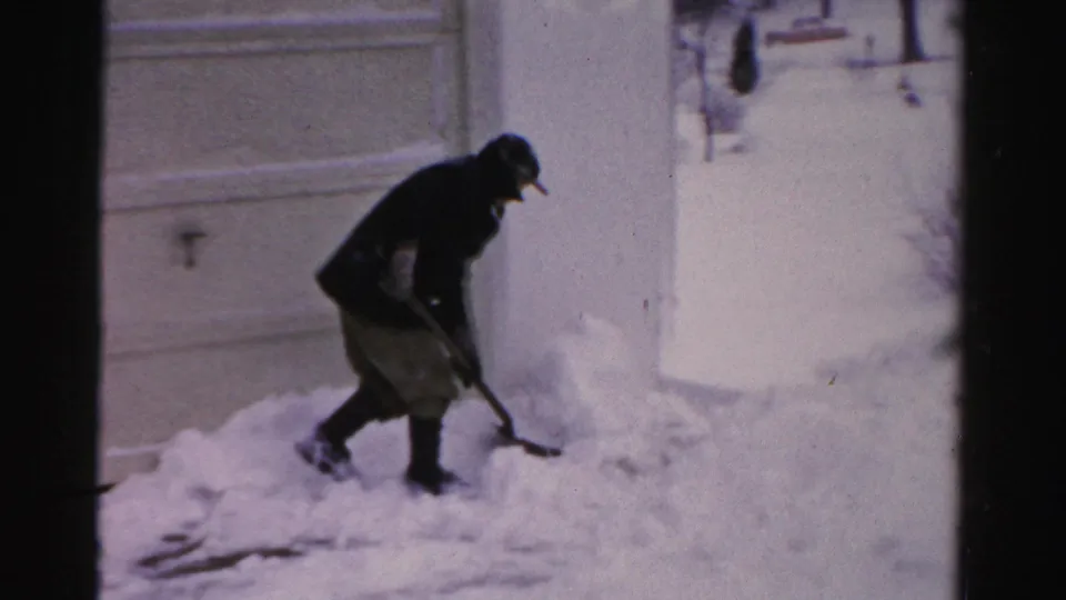 In Albany, New York home-movie footage, a man in a dark coat and cap bends to shovel snow near a residential garage or house wall.