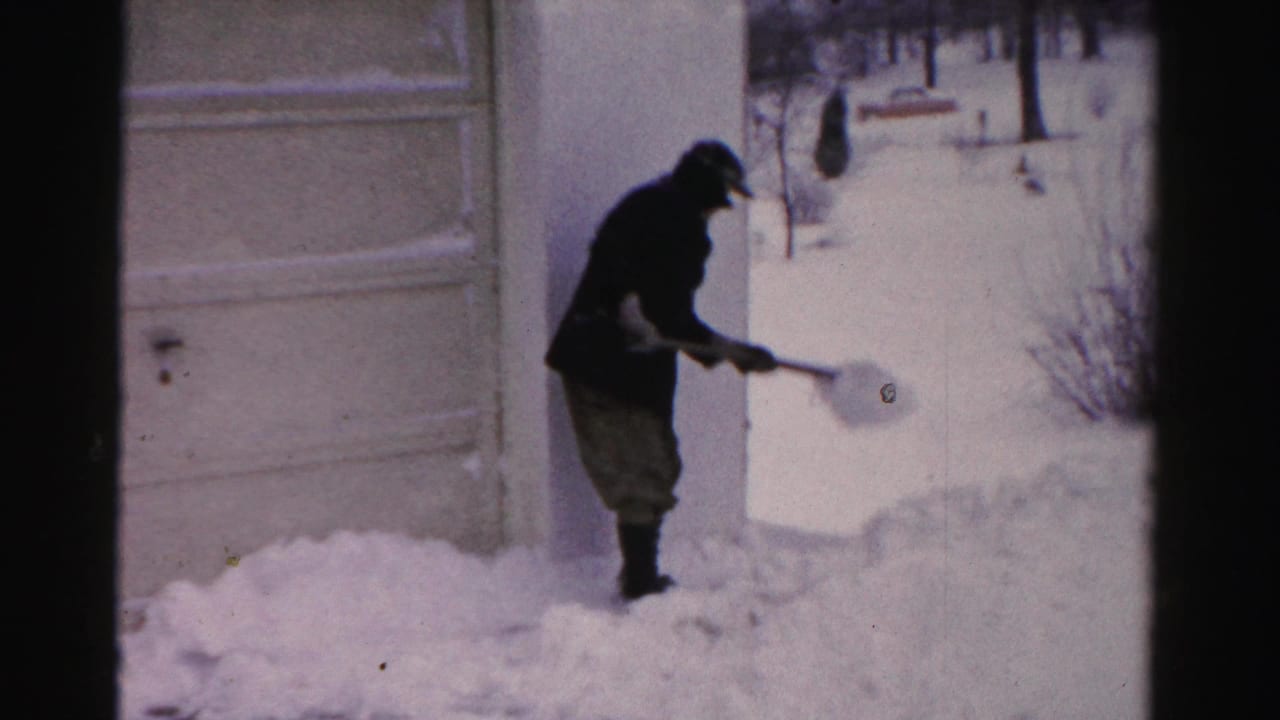 Man stands in snow and throws a shovelful near a residential driveway.