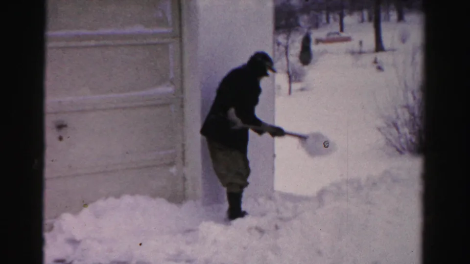 A man in winter clothing tosses snow with a shovel outside a house in Albany, New York, with deep snow covering the driveway area.