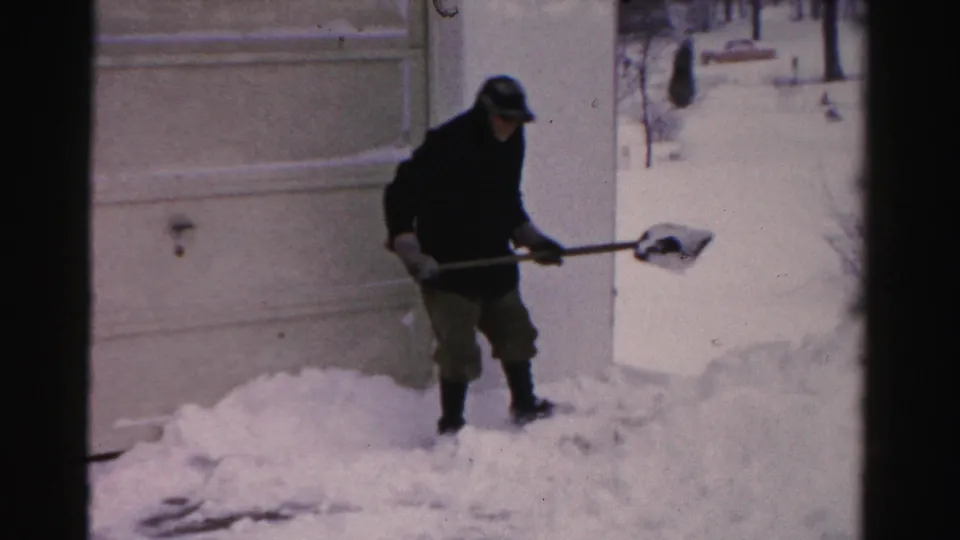 A man in dark winter clothing holds a shovelful of snow near a house exterior in the Albany, New York winter footage.