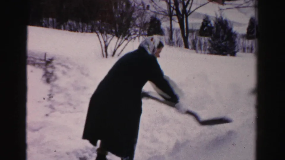 Another man, wearing a dark coat and light head covering, shovels beside a high snowbank in a snowy Albany residential yard.