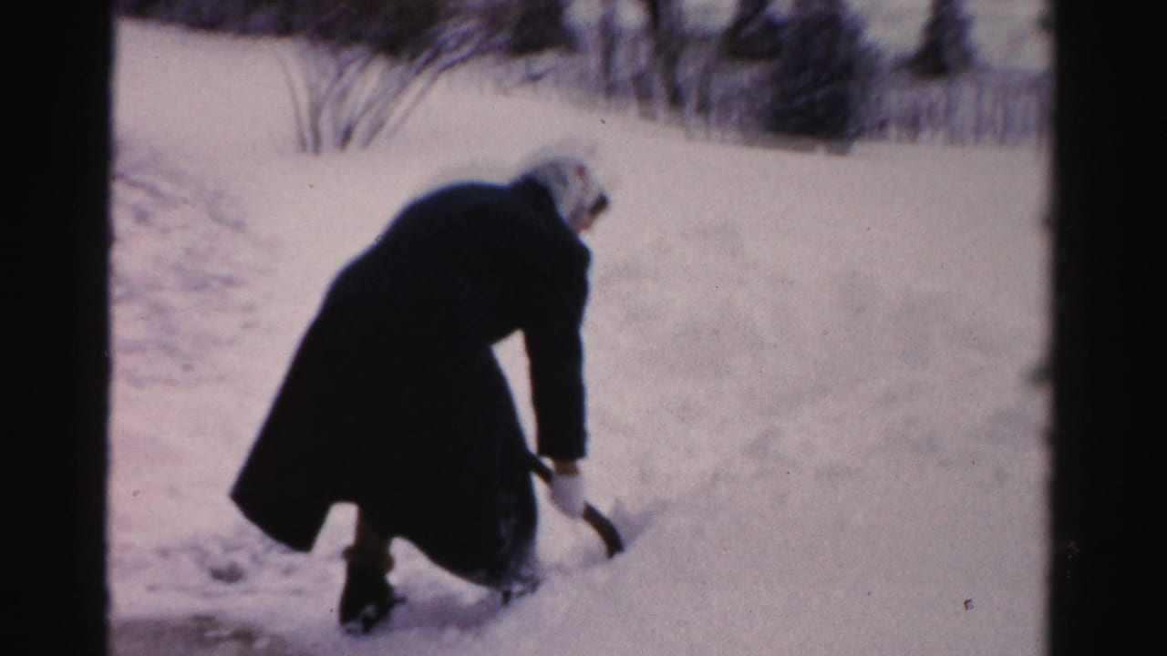 In Albany winter home-movie footage, a bundled person in a dark coat bends to shovel deep snow near a yard path.