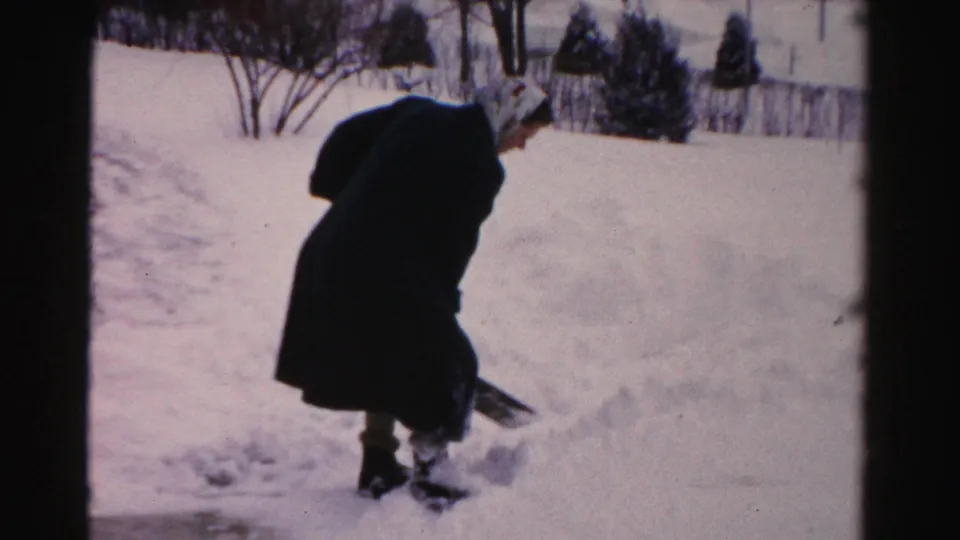 A bundled person works with a shovel in a snow-covered Albany yard, with bare trees and fence faintly visible behind.