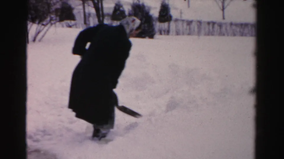A person in a long dark coat pauses with a shovel amid deep snow in the Albany residential yard scene.
