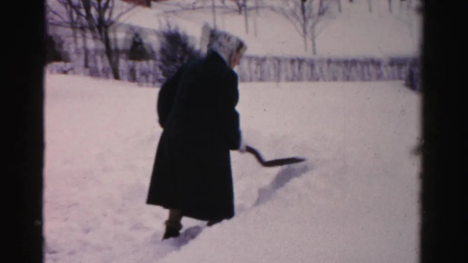 A bundled person stands beside a snowbank with shovel in hand during a 1950s Albany winter yard scene.