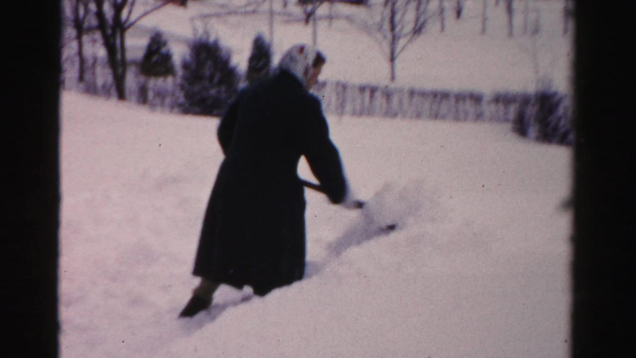 In Albany, New York home-movie footage, a woman in a dark coat and headscarf begins shoveling deep snow in a residential yard.