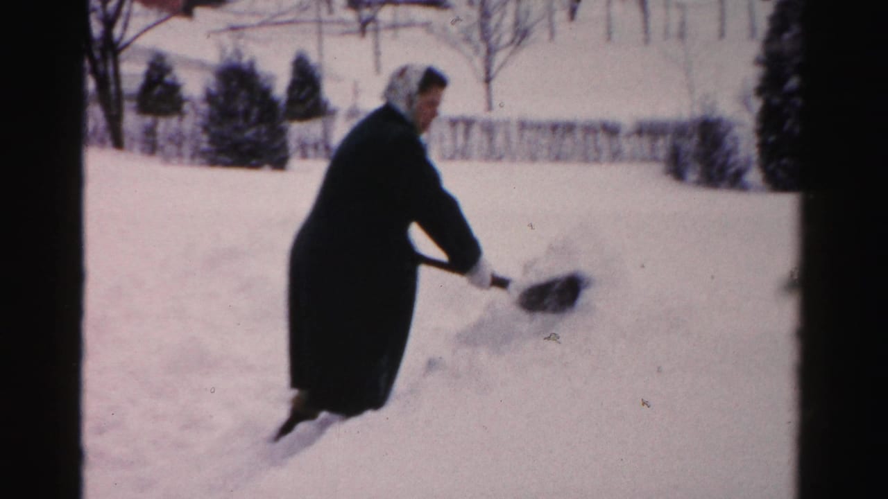 Woman pushes a shovel through snow with trees and fence behind her.