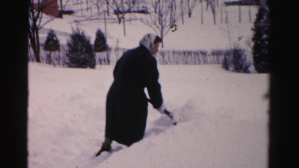 A woman in winter clothing bends over the shovel while clearing snow in a residential Albany yard captured on 8mm film.