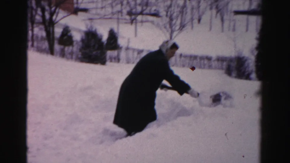 The woman extends the shovel to throw snow aside, with a low snowbank, bare trees, and fence line in the background.