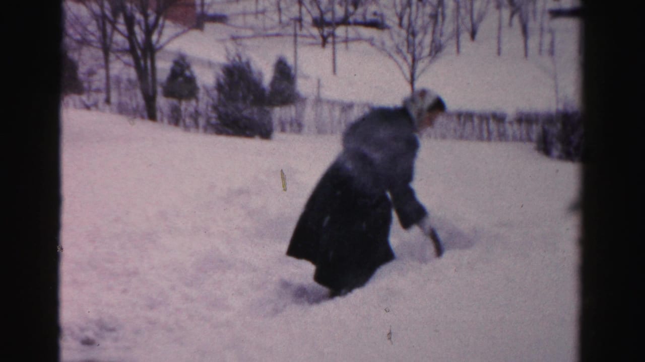 In Albany winter home-movie footage, a woman in a dark coat and headscarf works with a snow shovel in deep yard snow.