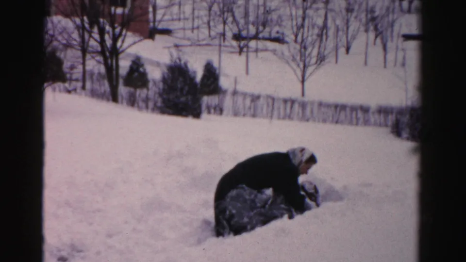 The woman bends over the shovel and gathers snow in the residential yard, framed by snowbanks and winter trees.
