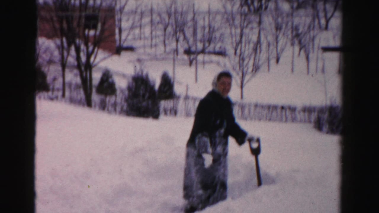 Woman stands with a shovel in deep snow, looking toward the camera in a snowy yard.