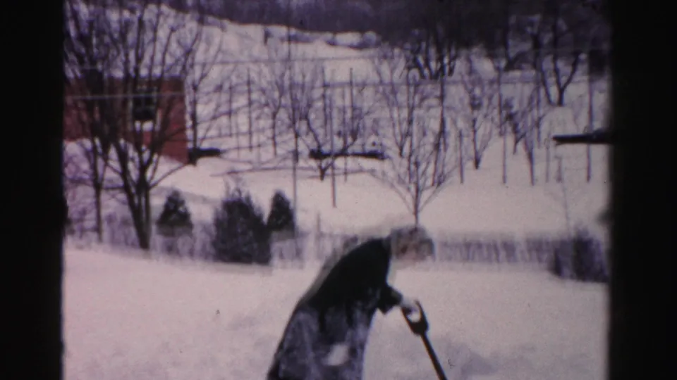 The Albany home-movie frame shows the woman bending into the task of shoveling snow near bare trees and a brick house.