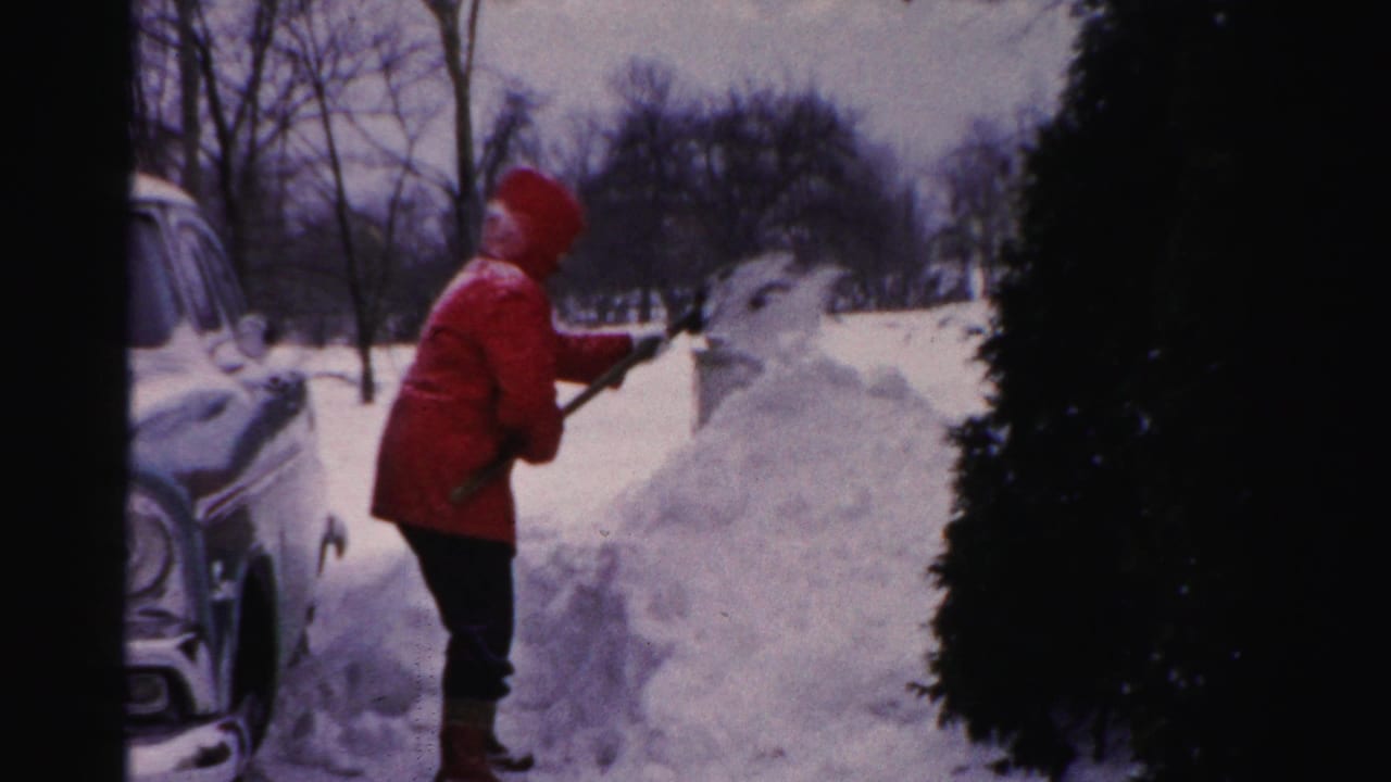 In Albany, New York, an adult in a red hooded coat shovels deep snow beside a 1950s car in a residential winter setting.