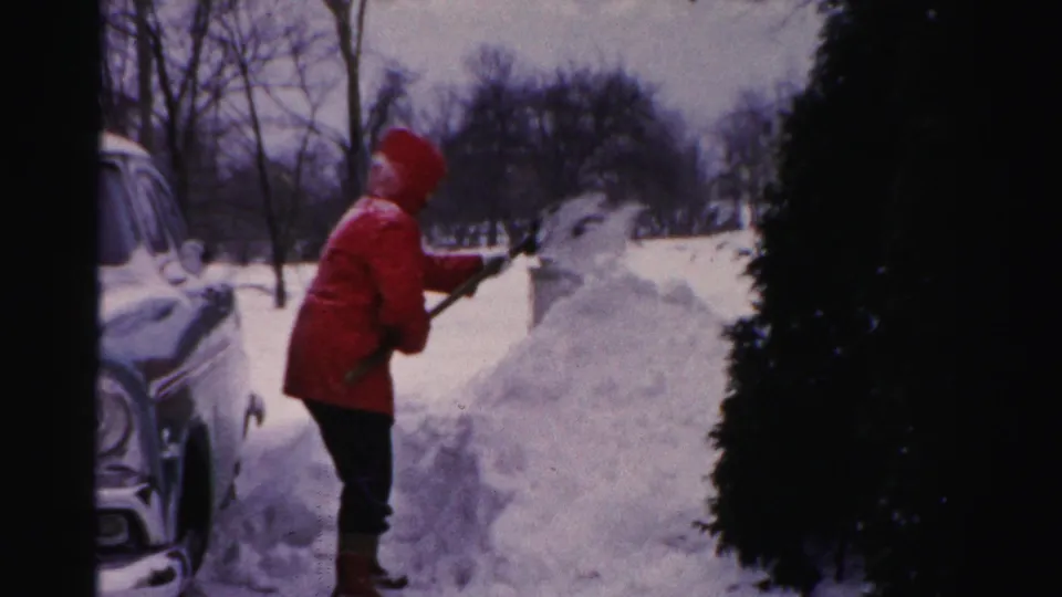 In Albany, New York, an adult in a red hooded coat shovels deep snow beside a 1950s car in a residential winter setting.