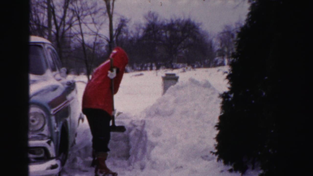 Adult in a red coat lifts a shovel near a snowbank and parked car.