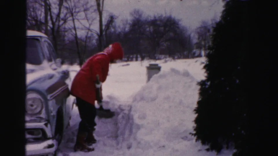 An adult in winter clothing bends over a shovel while clearing packed snow beside the car in the Albany home-movie scene.