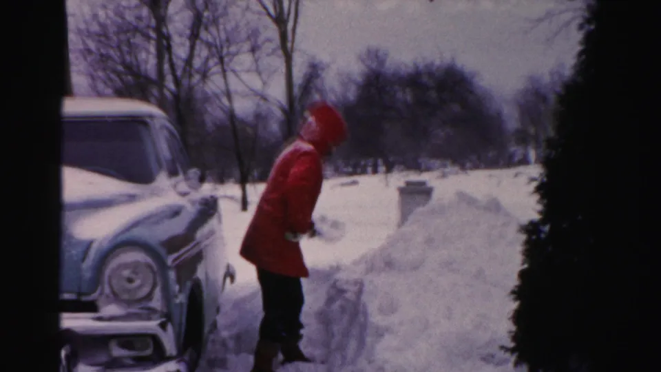 The adult stands near the shoveled snowbank, with the front of a 1950s car, bare trees, and an evergreen shrub framing the winter yard.