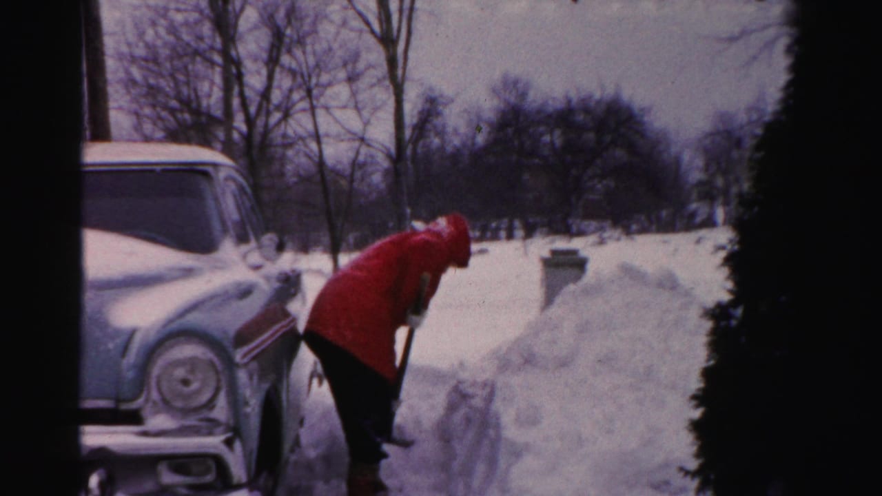 In Albany winter home-movie footage, a person in a red parka bends over a shovel beside a snow-covered vintage car.