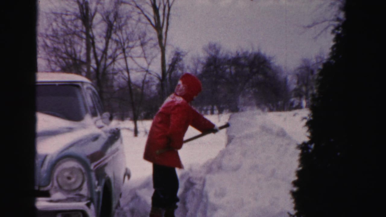 Person in red winter clothing lifting snow beside a vintage car and snowbank.