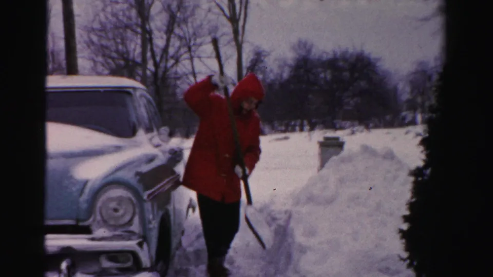 The red-coated figure stands near the car with a shovel, framed by snowbanks and leafless trees in the winter yard.