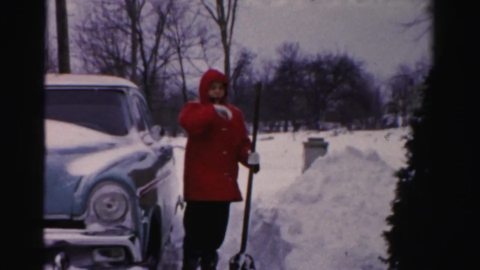 A person in a red coat holds a shovel near a plowed snow pile, with a vintage car partly visible at left.