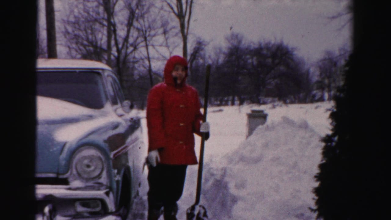 In Albany winter home-movie footage, a person in a red hooded coat holds a shovel beside a snow-covered mid-century sedan.