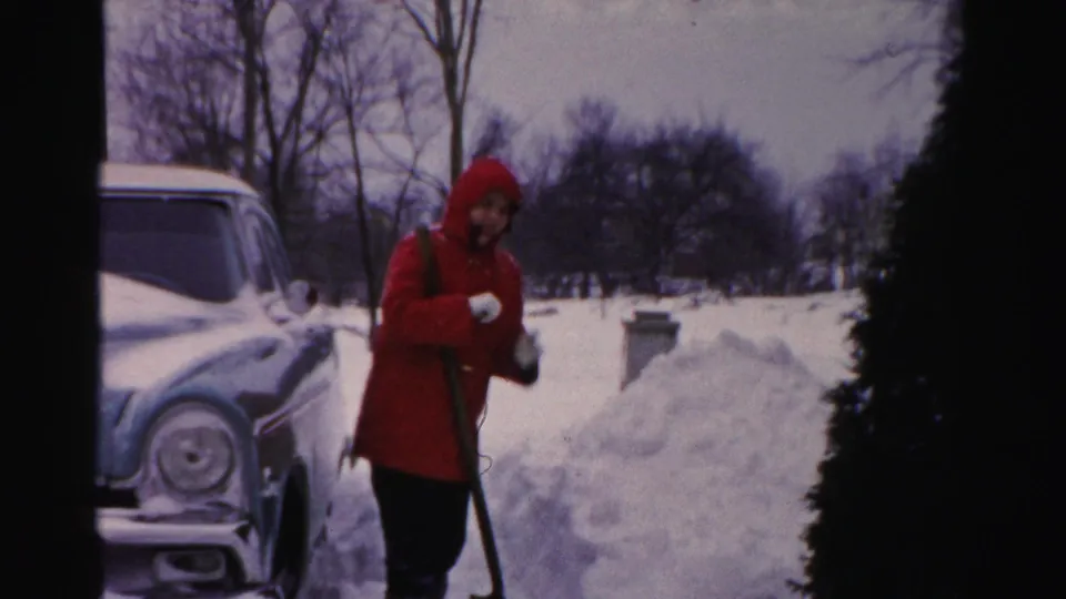 The Albany home-movie frame shows a person in a red hooded coat handling a shovel next to a snowy mid-century car.