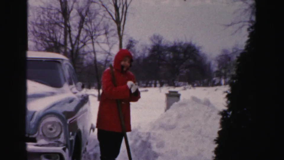 A person in a red coat appears to face or wave toward the camera while standing with a shovel in a snowy Albany yard.