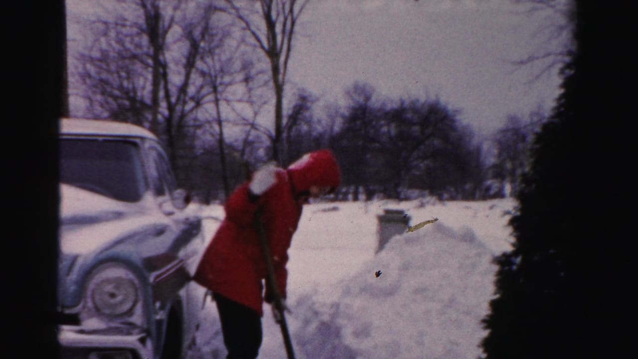 In Albany-area 1950s home-movie footage, a woman in a red hooded coat bends over a snow shovel beside a snow-covered blue car.