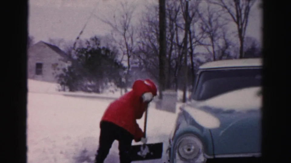 The woman leans into the shovel while clearing snow, with a snow-dusted blue car and bare winter trees in the background.