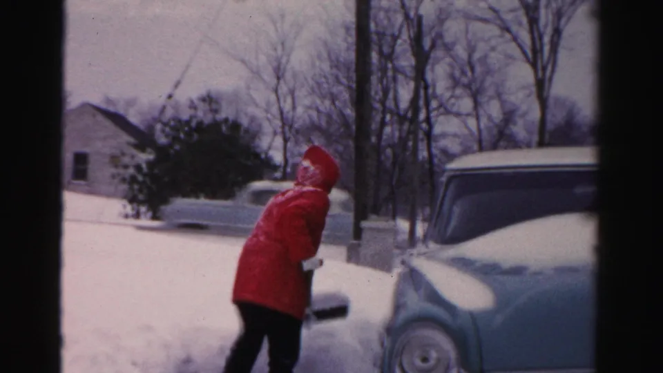A woman in a red hooded coat stands after shoveling near a pale blue car, with snowy ground, bare trees, and houses behind her.