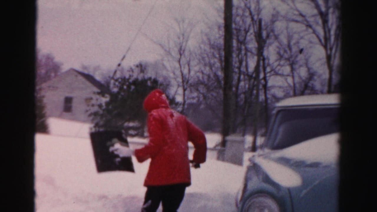 In Albany, New York home-movie footage, a person in a red hooded coat lifts snow near a pale blue parked car.